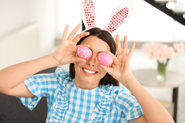 Beautiful woman in bunny ears headband holding Easter eggs near eyes at home