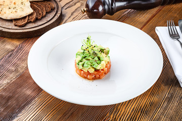 Close up view on fresh salmon tartare with avocado and microgreen in white plate. Homemade tartare on wooden background with copy space. Served food for menu or recipe. Italian cuisine snack