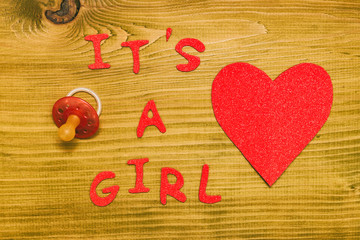 Text it's a girl with pacifier and heart shape on wooden table.Toned photo.