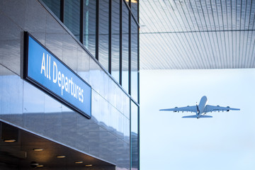 all departures sign at airport building with aircraft taking off in background