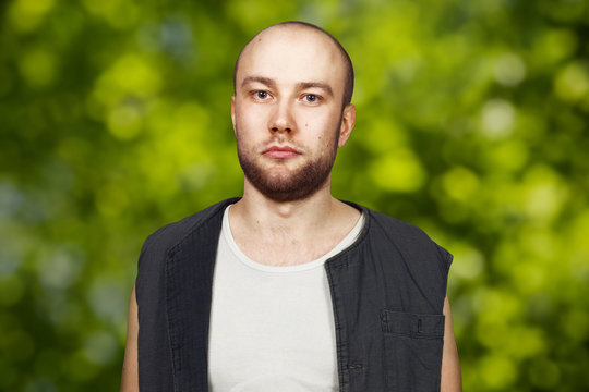 Portrait Young bald guy with beard with pocker face dressed in sleeveless shirt. Man on green bokeh background.