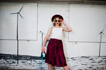 Obraz premium Fashionable and beautiful blonde model girl in stylish red velvet velour skirt, white blouse, hat and sunglasses, posed with phone and earphones.