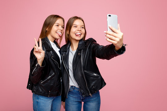 Gorgeous two young twin sisters in leather jackets with bright smile use smartphone over pink background. Twin sisters take selfie with mobile phone.