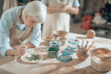 Cropped photo of elderly craftswoman painting decorative plate in workshop
