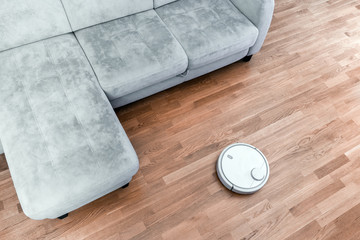 Obraz na płótnie Canvas Robotic vacuum cleaner on laminate floor near sofa closeup, smart home robotics wireless cleaning for simplify routine housework, efficient dust absorption in absence of householder