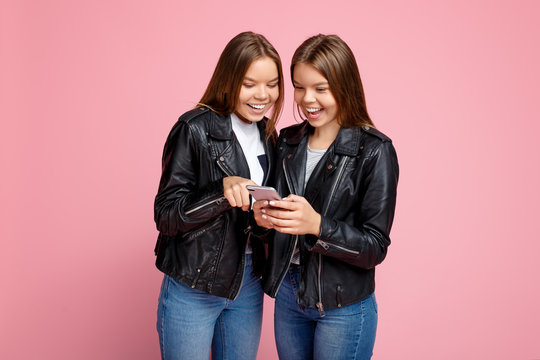 Funny two young twin sisters with bright smile in leather jackets use smartphone over pink background.