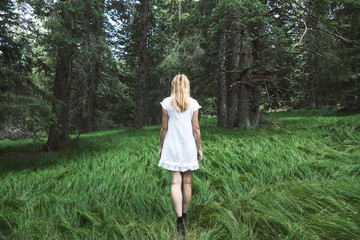 Single woman in white dress walks in dreamy green forest fairytale.