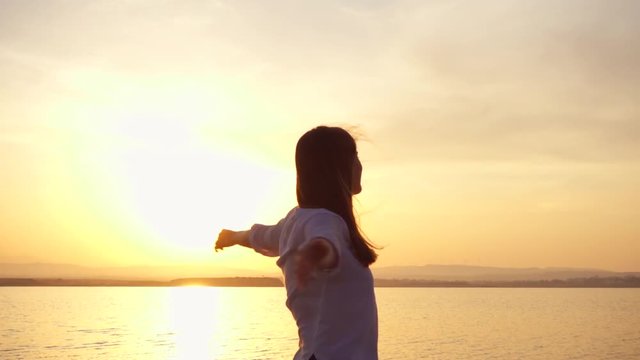 Dark silhouette of young woman raising arms up and dancing at sunset on lake. Female figure outstretching hands at golden hour in slow motion