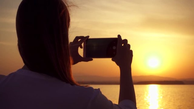 Dark silhouette of young woman taking photos on cellphone at sunset on lake while enjoying quiet moment in nature. Carefree female figure making pictures on mobile phone at golden hour in slow motion