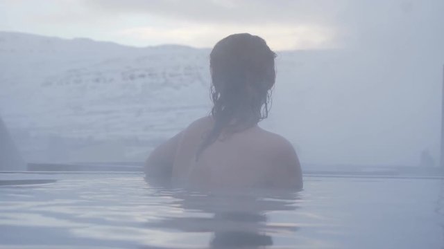 Back view of young girl in open hot thermal swimming pool. Traveling Pretty young lady relaxing in mountains valley. Enjoy in pool at Winter day