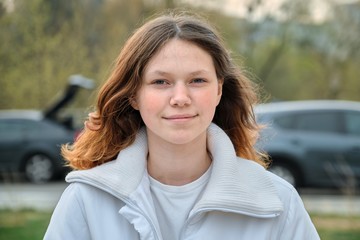 Outdoor portrait of teenager girl 15 years old, girl smiling with long brown hair in white jacket