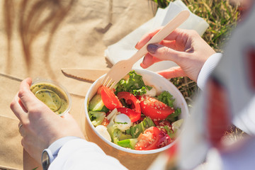Woman eating healthy salad from paper container over green grass sitting in local park on a sunny...