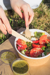 Woman eating healthy salad from paper container over green grass with flying hair on a sunny spring day. backlight