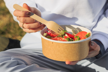 Woman eating healthy salad from paper container over green grass sitting in local park on a sunny spring or summer day, sunlight