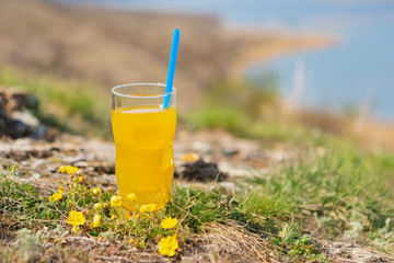 a glass with yellow fresh orange juice and with a blue straw, stands against a beautiful landscape, the concept of travel and healthy lifestyle
