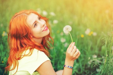 Portrait of young redhair woman blowing dandelion with joy in spring garden. Springtime Beautiful girl at sunset in meadow landscape. Allergic to pollen of flowers. Spring allergy free healthy concept