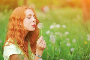Portrait of young redhair woman blowing dandelion with joy in spring garden. Springtime Beautiful girl at sunset in meadow landscape. Allergic to pollen of flowers. Spring allergy free healthy concept