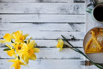 A cup of tea and pancakes on a plate. Yellow daffodils on a white wooden table.