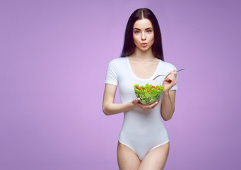 Beautiful slim brunette young girl in white lingerie on purple background. Sporty healthy model holding fresh vegetable salad bowl. Sport, diet, healthy food, nutrition, lifestyle, fat burn concept.