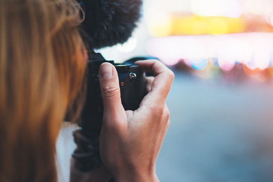 hipster girl hold in hands modern photo camera, take photography click on defocus background bokeh light in evening street attraction, photographer look on camera technology, night city illumination