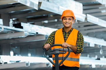 smiling indian warehouse worker standing near pallet jack, smiling and looking at camera