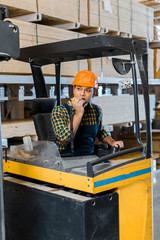 Fototapeta na wymiar handsome indian warehouse worker sitting in forklift machine and talking on walkie talkie