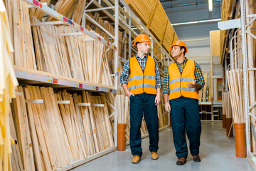 Fototapeta premium smiling multicultural workers talking while walking along racks with wooden construction materials