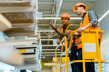 serious indian worker pointing with hand at shelf with construction materials while standing on scissor lift with colleague
