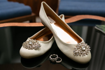 Womens white ballet flats with a brooch on a sock and rings lying on a glass table. © melnikviva