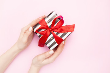 woman hands with Gift present box in white and black stripes with a red ribbon bow isolated on pink table top view. Minimal flat lay composition for sales, birthday, mother day or christmas. copyspace