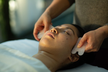 Young woman receives facial rejuvenation with gua sha rose quartz scraping in spa wellness center