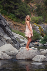 A young, barefoot woman in a summer dress standing on a rock in a mountain creek.
