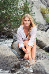 Young blond woman sitting on a rock with her feet in a mountain creek after a hike.