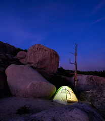 Illuminated tent on a camping trip at night on a barren, rocky landscape in the wilderness surrounded by granite boulders.