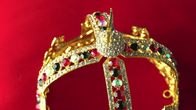 Gold crown with luxury color jewels and diamonds, view from above to the middle of the frame. With reflection and lights sparks. Slow rotation on the red royal color surface. 4k, uhd. Macro close up.