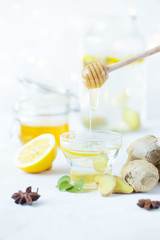 Honey is poured into a ginger drink in a cup. Ginger root, honey in a jar, lemon on a white table.