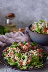 Taboule with pearl barley porridge with fresh vegetables and herbs on a plate and bowl on a gray concrete background
