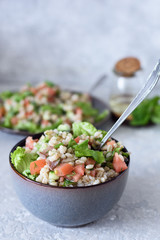 Taboule with pearl barley porridge with fresh vegetables and herbs on a plate and bowl with a spoon on a gray concrete background