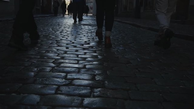 People's legs walking along pavement with sidewalk tiles in city.