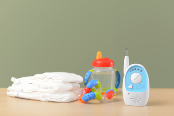 Modern baby monitor with toy, bottle and diapers on table against color background