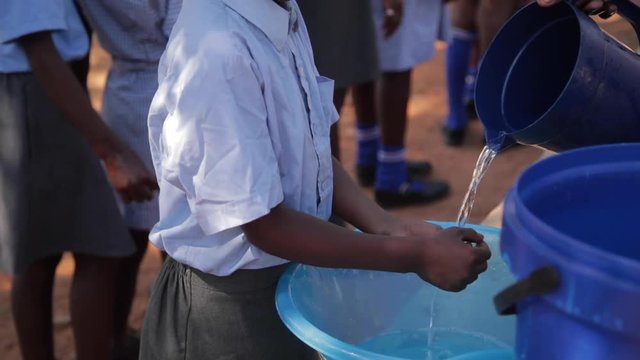 African school pupil steps up and washes his hands in a plastic dish with water poured from a jug outside before mealtime