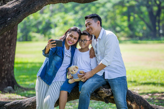 happy asian Family, parents and their children taking selfie in park together. father, mother ,son sitting on branch of big tree take a photo by smart phone having fun and laughing outside .cheerful