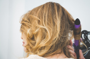 Fototapeta premium Beuty and fashion concept, Hairdresser curling woman hair with electric iron curler tong, blonde hair