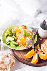 Breakfast eggs on a white plate with half avocado, cucumbers on a wooden plate on bed