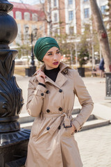 Stylish Muslim girl with a scarf on her head in a beige raincoat in the city. Stylish headband. The image of a modern woman in the city on the background of architecture