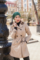 Stylish Muslim girl with a scarf on her head in a beige raincoat in the city. Stylish headband. The image of a modern woman in the city on the background of architecture