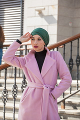 Stylish Muslim girl with a scarf on her head in a pink coat in the city. Stylish headband. The image of a modern woman in the city on the background of architecture