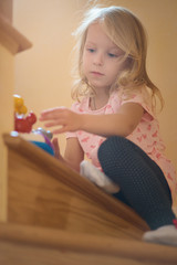 Fototapeta premium little girl sits on the stairs to the second floor of the house, playing lonely. sunlight from the window illuminates. concept of family, childhood