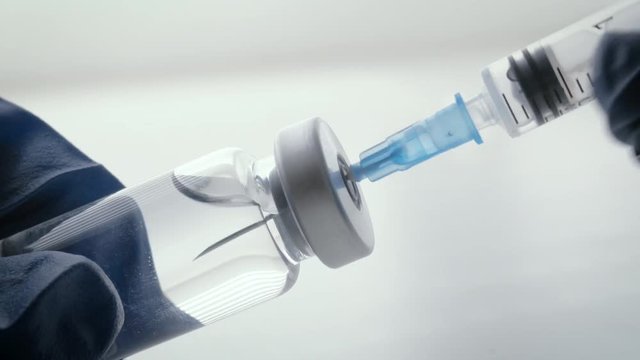 Filling syringe with mandatory vaccinations. A blue, gloved hand inserts the needle of the injection device into the rubber stopper of the vaccine vial and draws in the immunization.