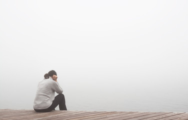 Junge Frau sitzt einsam am Rand eines Holzweges einer Brücke gebückt und traurig in Gedanken.